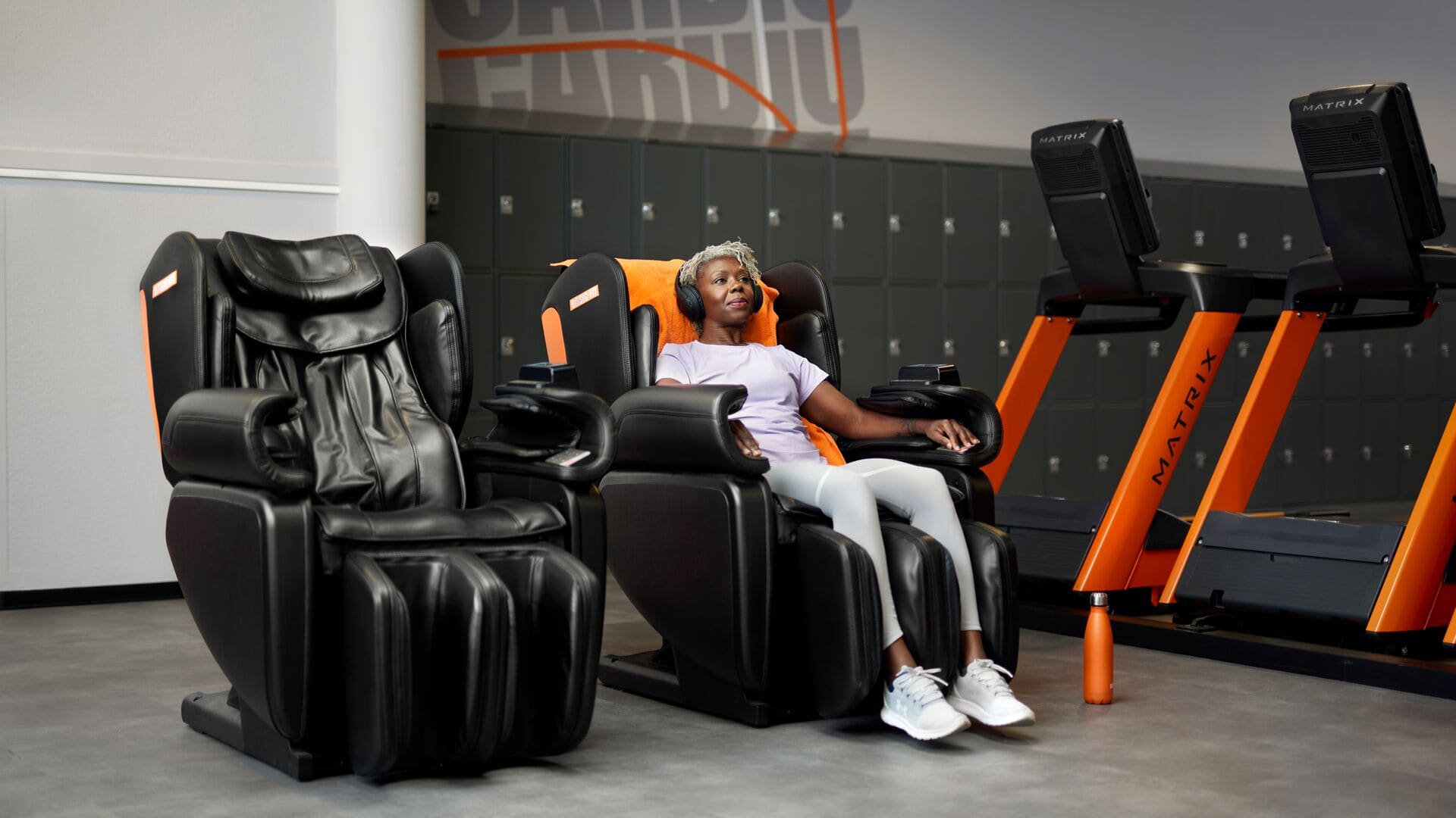 Een vrouw ontspant in een zwarte massagestoel met oranje accenten, met een koptelefoon op, in een Basic-Fit club. Ze zit comfortabel na haar workout, met loopbanden en lockers op de achtergrond.