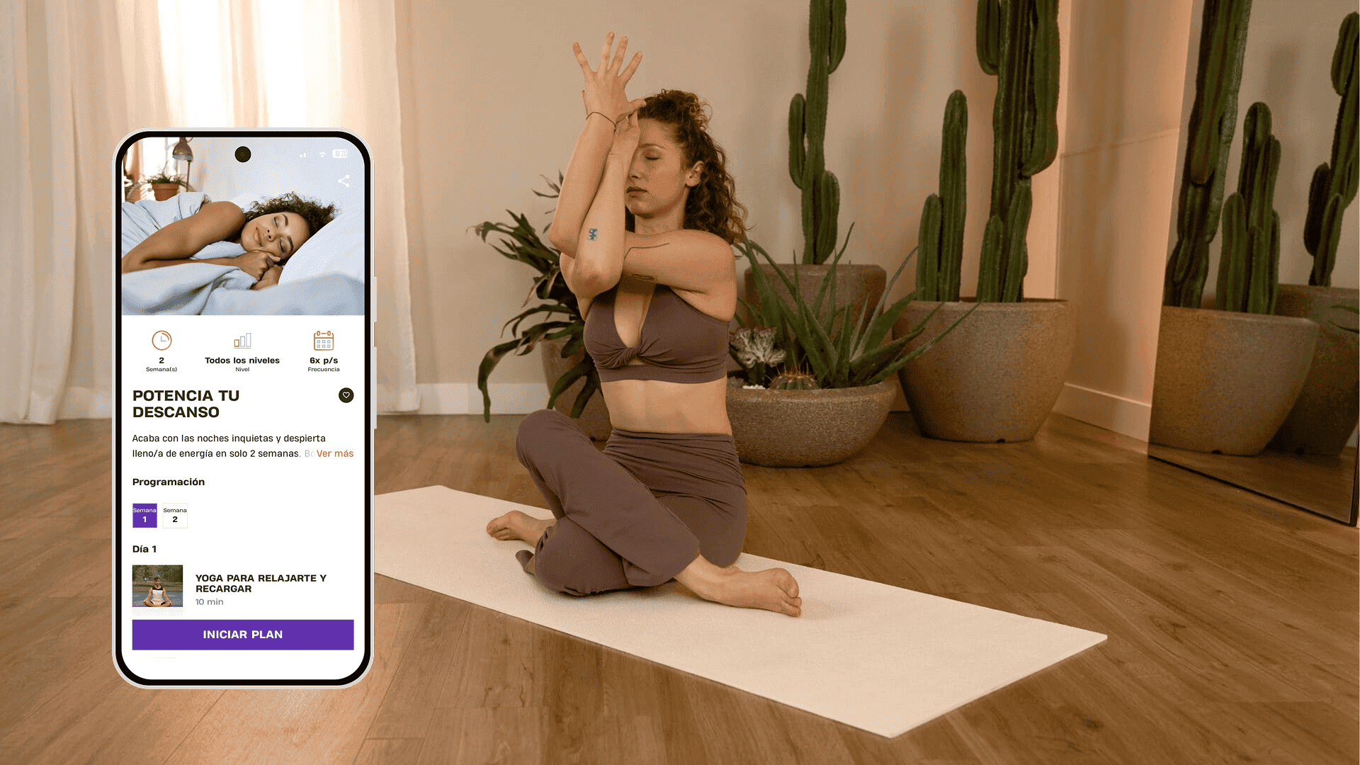 Una mujer practica yoga sobre una esterilla en una habitación cálida y tranquila decorada con grandes plantas. Está sentada con las piernas cruzadas y se concentra en un estiramiento de brazos y hombros. A su lado, un teléfono muestra el programa de Basic-Fit “Potencia tu descanso”, con sesiones de yoga para relajarse y recargar energía.