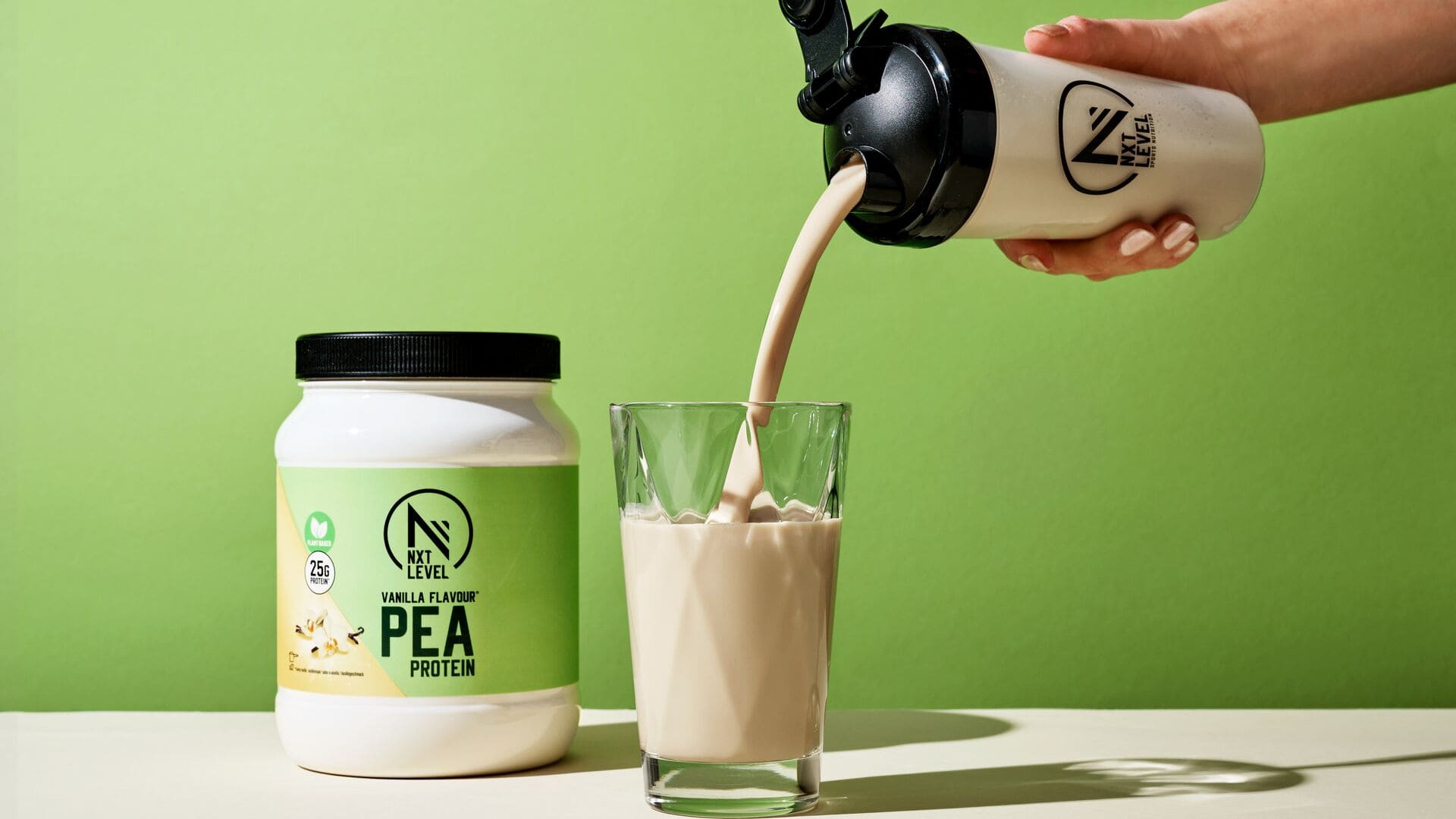 Main versant un shake protéiné aux pois saveur vanille dans un verre, à côté d’un pot de protéine NXT Level sur fond vert.