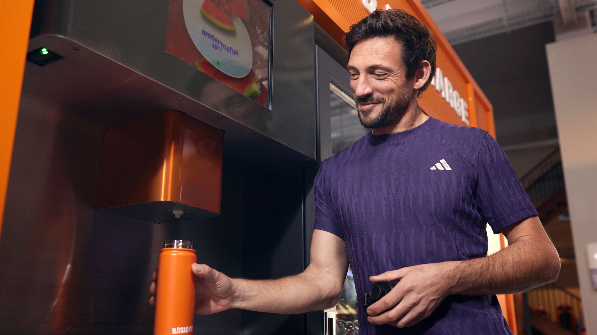 Een man vult een oranje Basic-Fit bidon bij een selfservice drankstation in een Basic-Fit club.