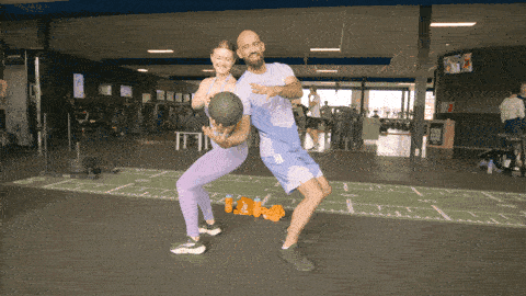 Two people standing back to back rotating to pass a medicine ball to each other, working on core rotation and coordination.