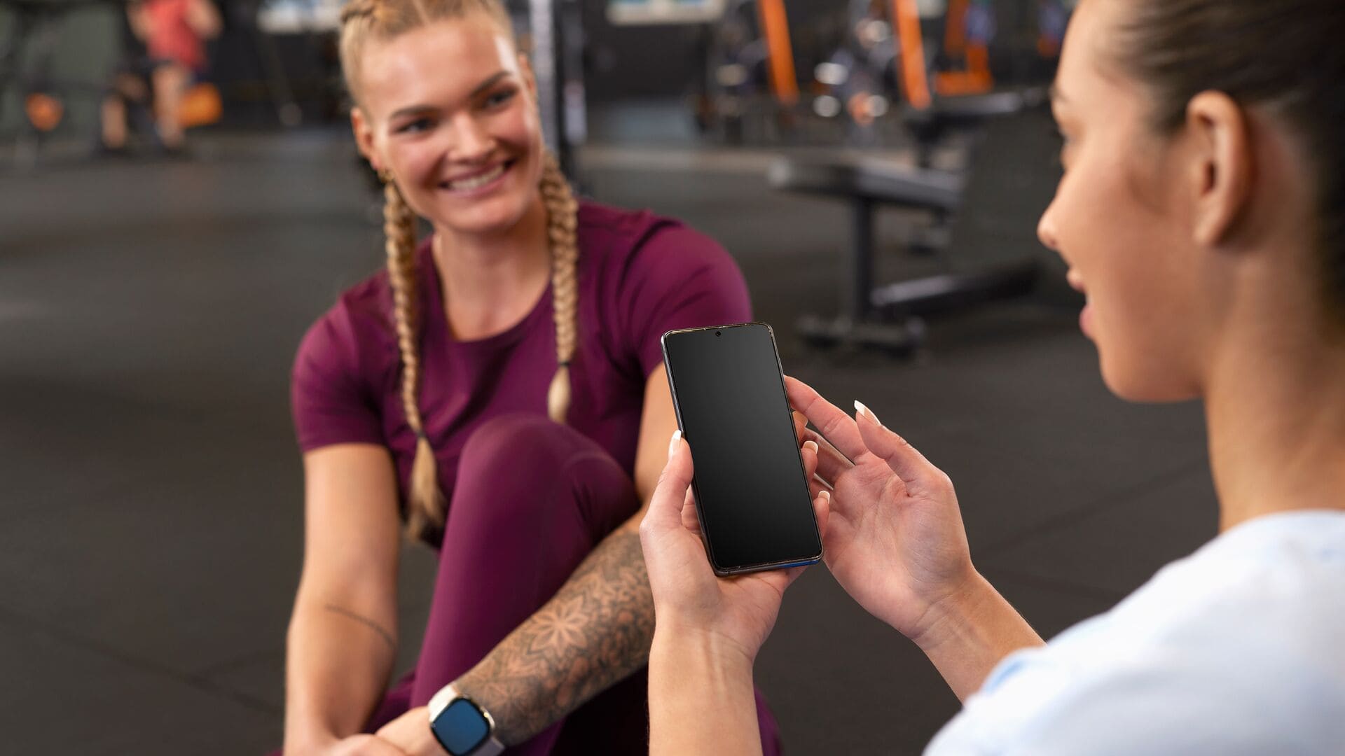 Zwei Frauen sitzen nach dem Training auf dem Boden im Fitnessstudio; eine hält ein Smartphone hoch, während die andere lächelt, beide in Sportkleidung.