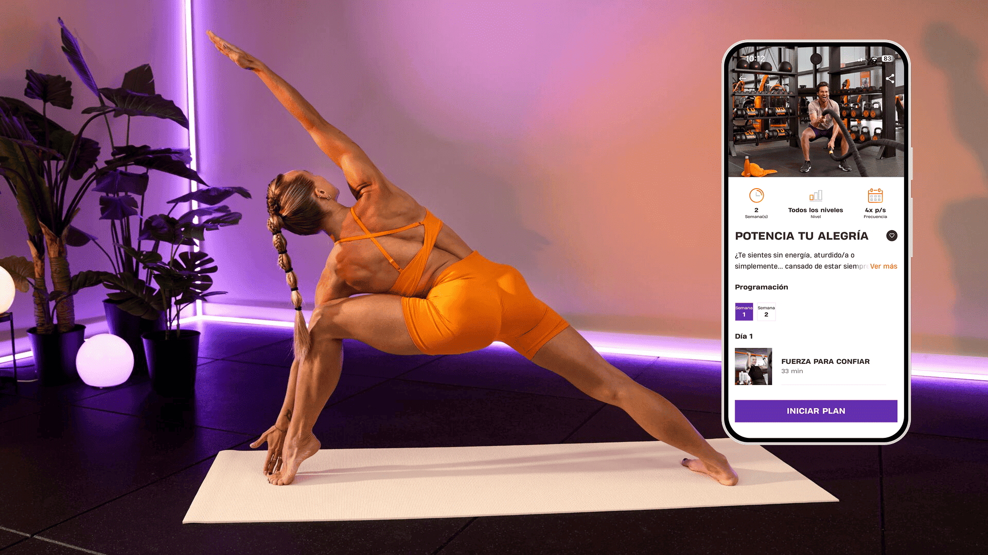 Una mujer realiza una postura de yoga sobre una esterilla en una habitación iluminada con luces moradas y decorada con plantas tropicales. Lleva ropa deportiva naranja brillante. A su lado, un teléfono muestra el programa de Basic-Fit “Potencia tu alegría”, con entrenamientos para ganar fuerza y confianza.