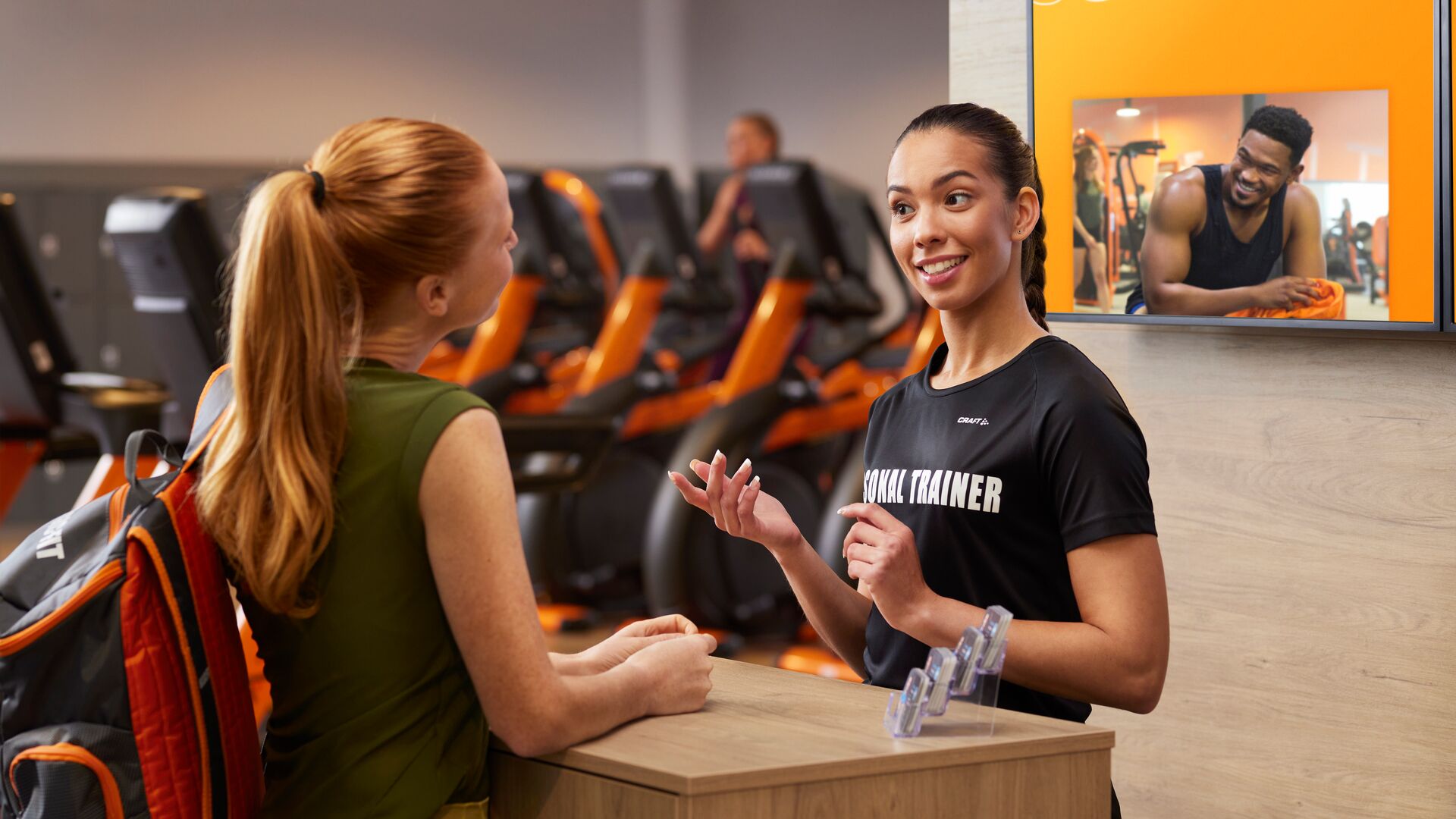Personal trainer bij een Basic-Fit club heet een lid vriendelijk welkom aan de balie, met oranje cardioapparaten en een digitaal scherm op de achtergrond.