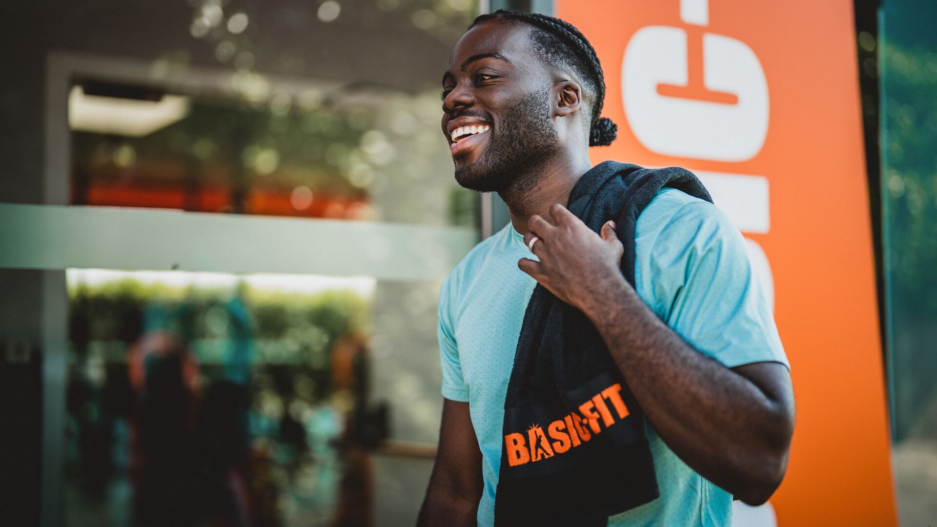 Smiling man in a light blue shirt standing outside a Basic-Fit gym, carrying a black towel with the orange Basic-Fit logo over his shoulder.