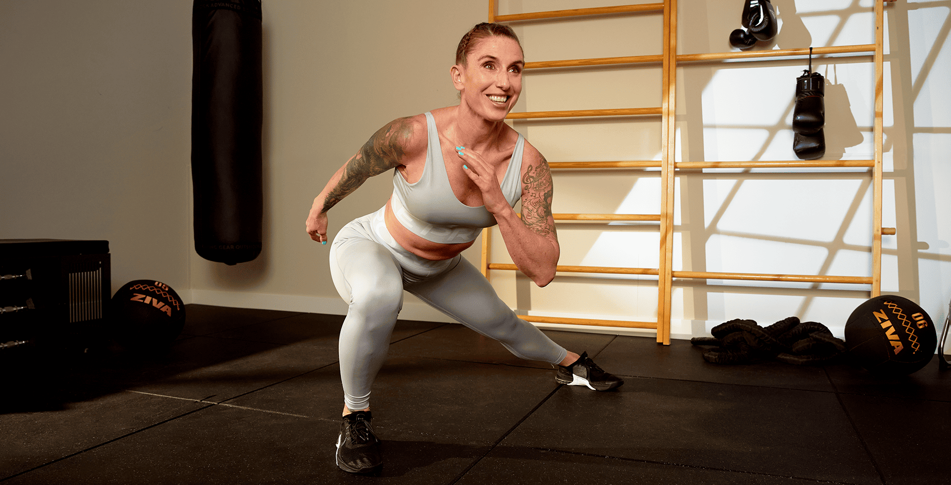 Eine Frau in heller Sportkleidung lächelt, während sie einen dynamischen Seitwärtsschritt in einem Basic-Fit-Trainingsbereich ausführt, umgeben von funktionellem Trainingsequipment wie einem Boxsack, Medizinbällen und Battle Ropes.