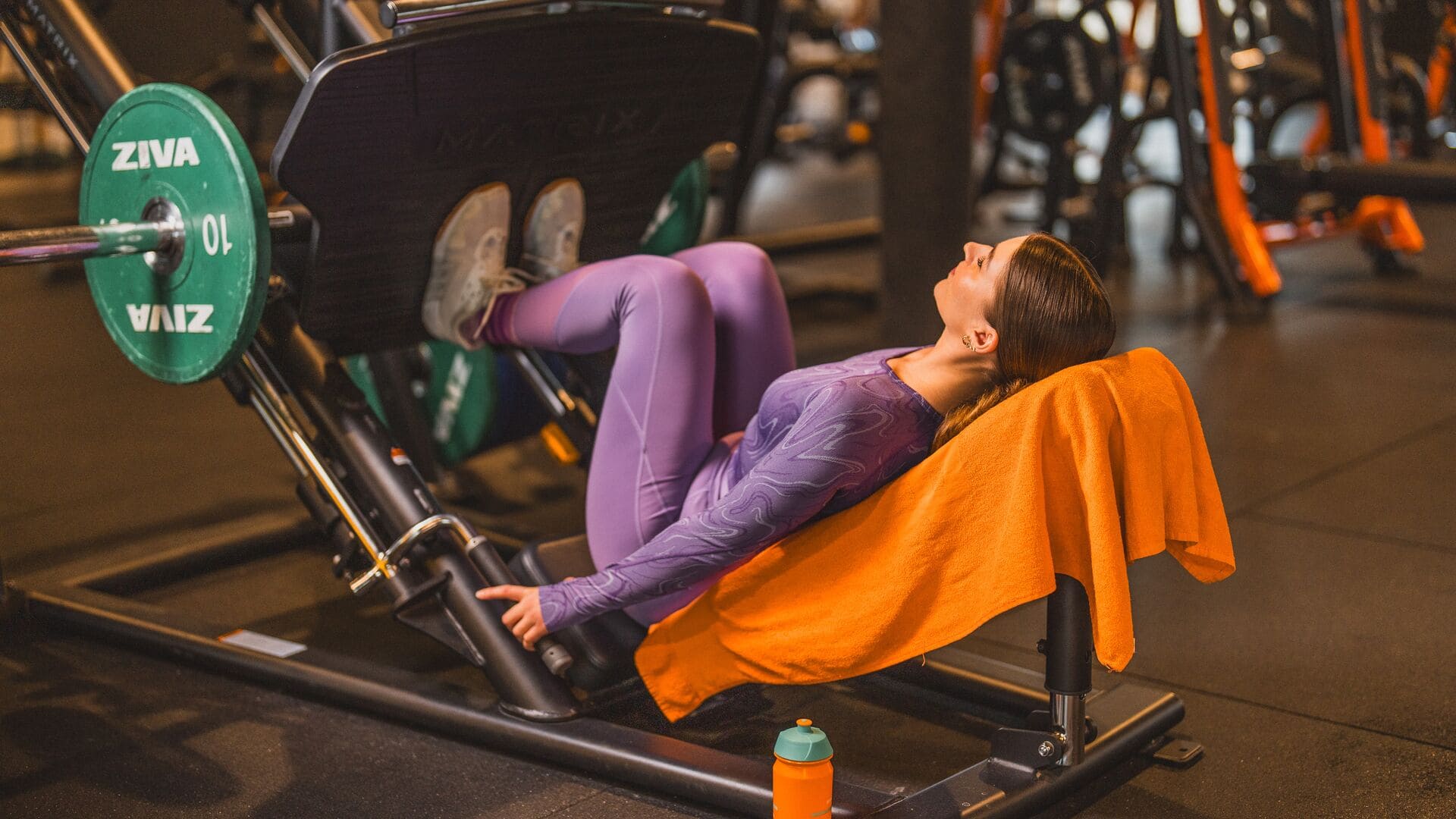 Eine Frau in violetter Sportkleidung trainiert an einer Beinpresse in einem Basic-Fit-Studio, mit einem orangefarbenen Handtuch auf der Bank und Gewichten daneben.