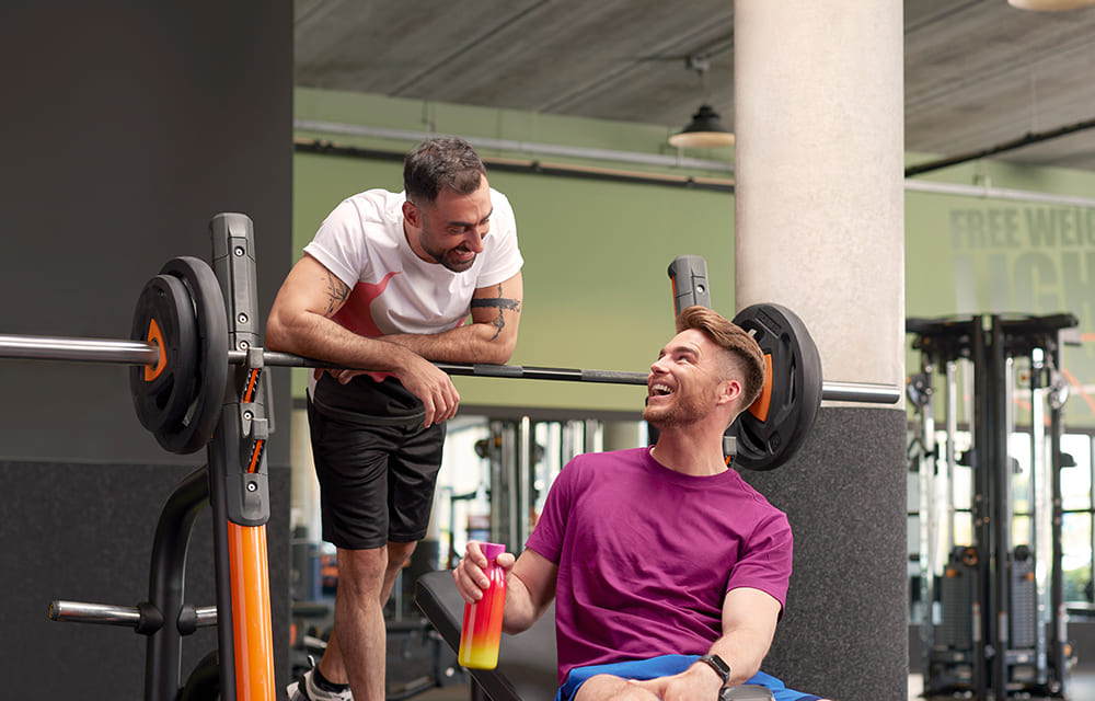 Zwei Männer befinden sich in einem Basic-Fit-Fitnessstudio, unterhalten sich und lachen neben einer Hantel auf einem Rack. Einer trägt ein weiß-rotes T-Shirt, der andere sitzt in einem violetten T-Shirt und hält einen Shaker in der Hand.