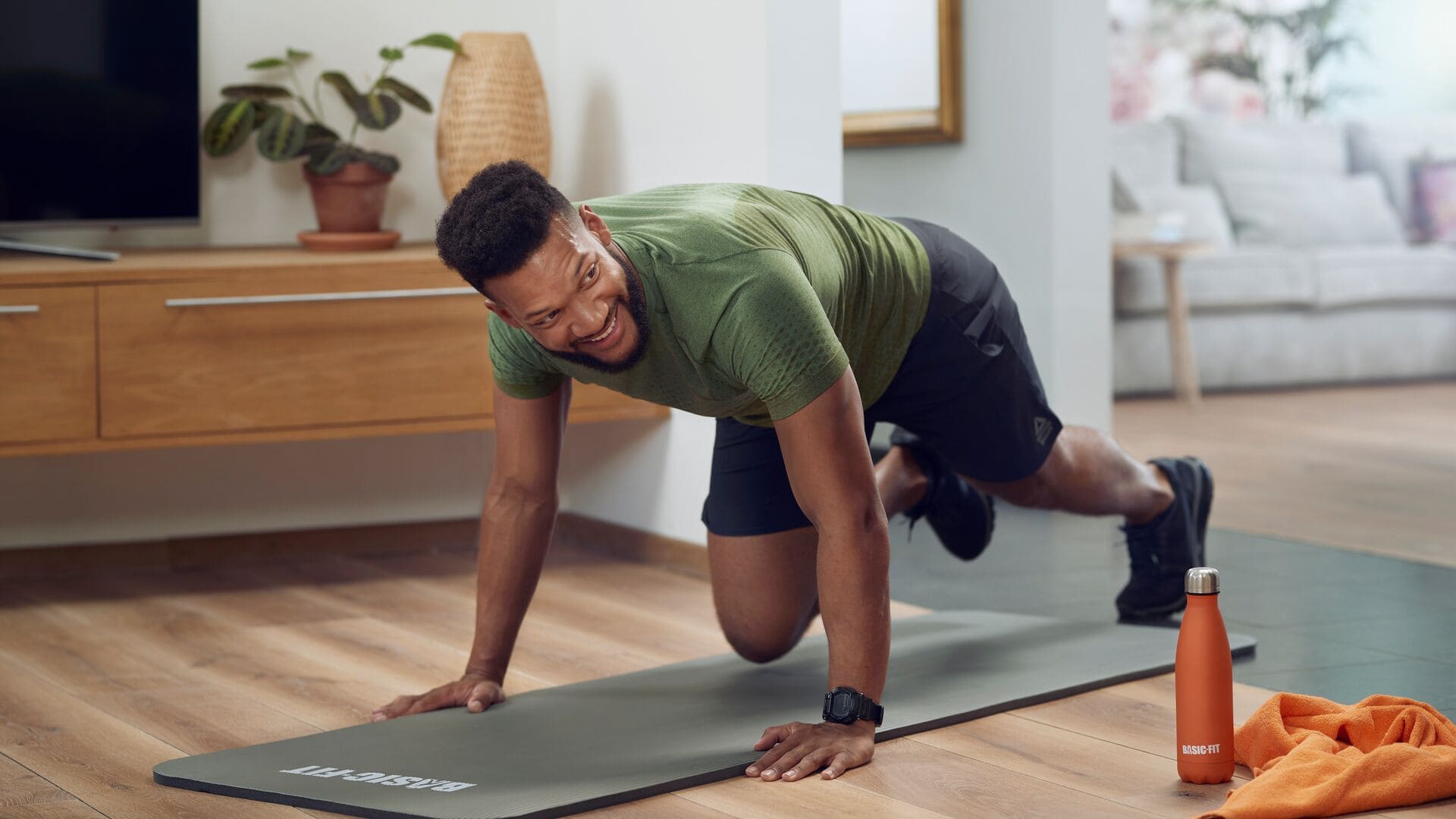 Ein Mann mit grünem T-Shirt und schwarzer Shorts trainiert zu Hause auf einer grauen Basic-Fit-Matte. Er lächelt, während er eine Plank-Position hält. Eine orangefarbene Basic-Fit-Trinkflasche und ein Handtuch liegen neben ihm auf dem Boden.