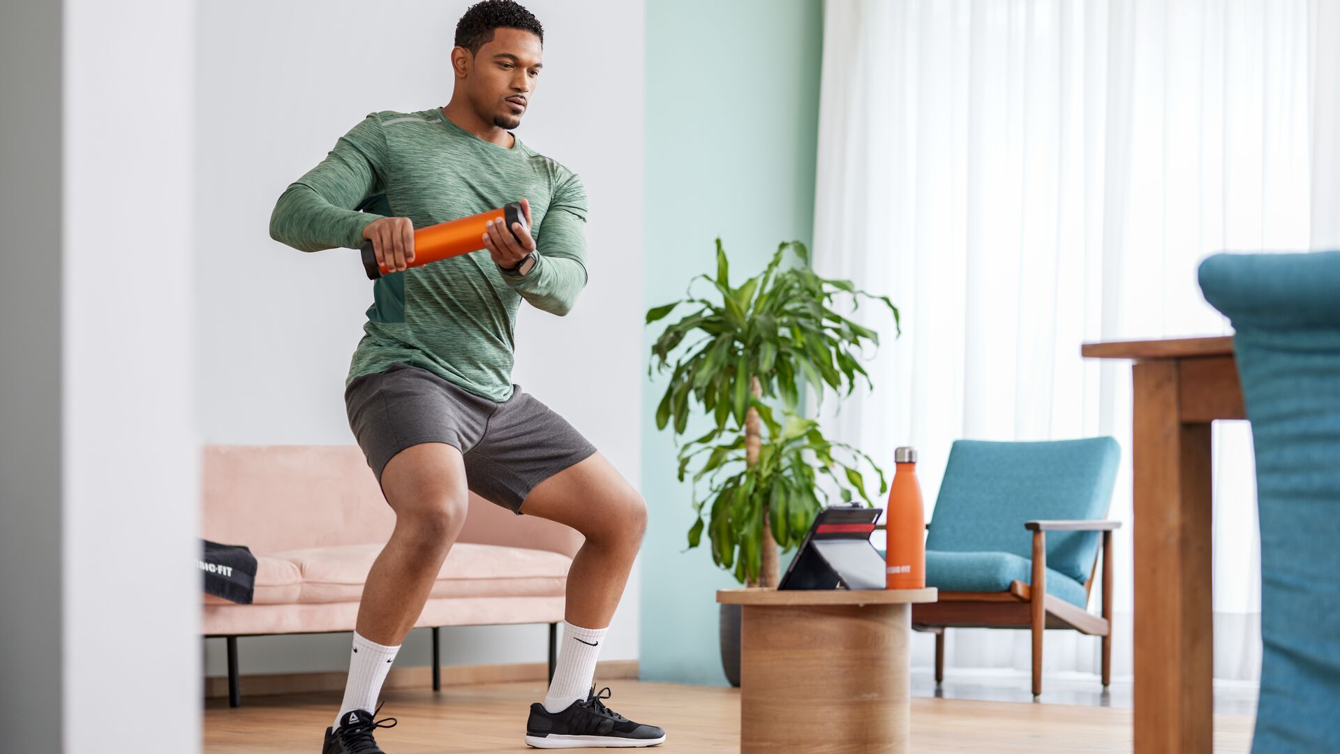Man exercising at home with an orange fitness tube, standing in a squat position. A tablet, water bottle, and indoor plants are visible in a bright living room with wooden furniture and light curtains.