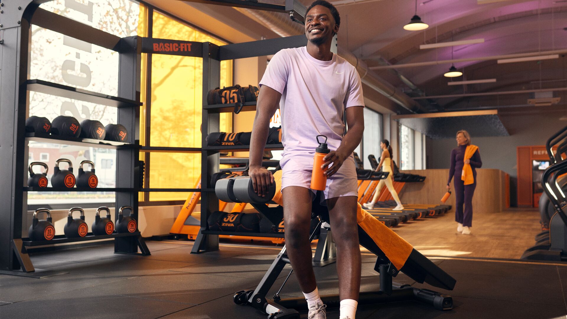 Person resting on a workout bench holding a water bottle in a gym, with kettlebells and cardio machines visible around the training area.