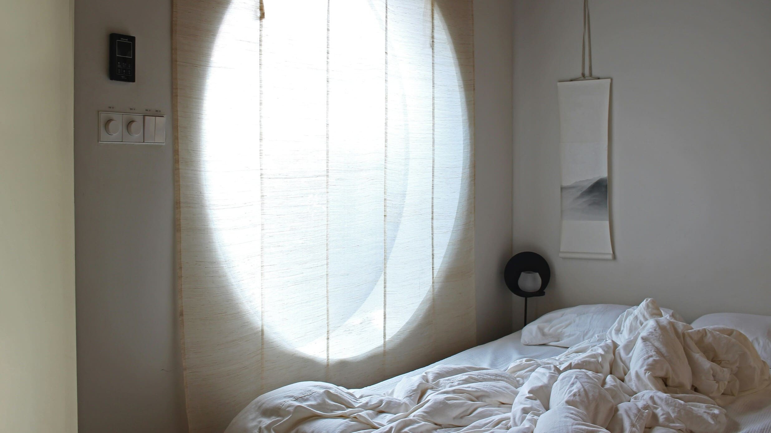 Sanftes Morgenlicht fällt durch leichte Vorhänge in ein ruhiges Schlafzimmer mit neutralen Farbtönen und einem ungemachten Bett.