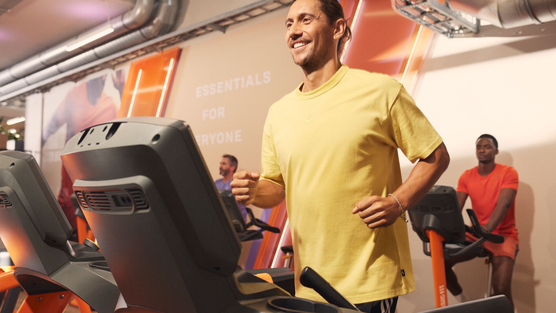 Een man loopt op een loopband in een Basic-Fit club en draagt een geel shirt, terwijl andere sporters achter hem trainen op cardioapparatuur in een helder verlichte ruimte met oranje details.