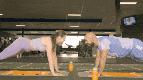 Two people in a high plank facing each other, tapping opposite hands in alternating reps to engage core and shoulders.