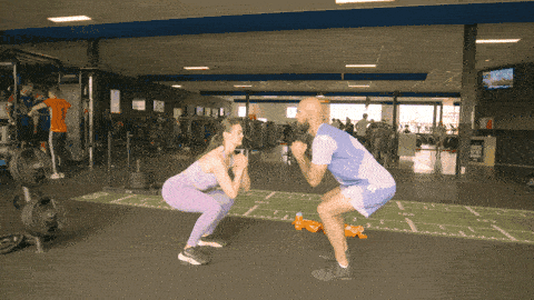 Two people side by side in a gym, one holding a squat while the other performs squats, then switching roles to alternate movement and static hold.