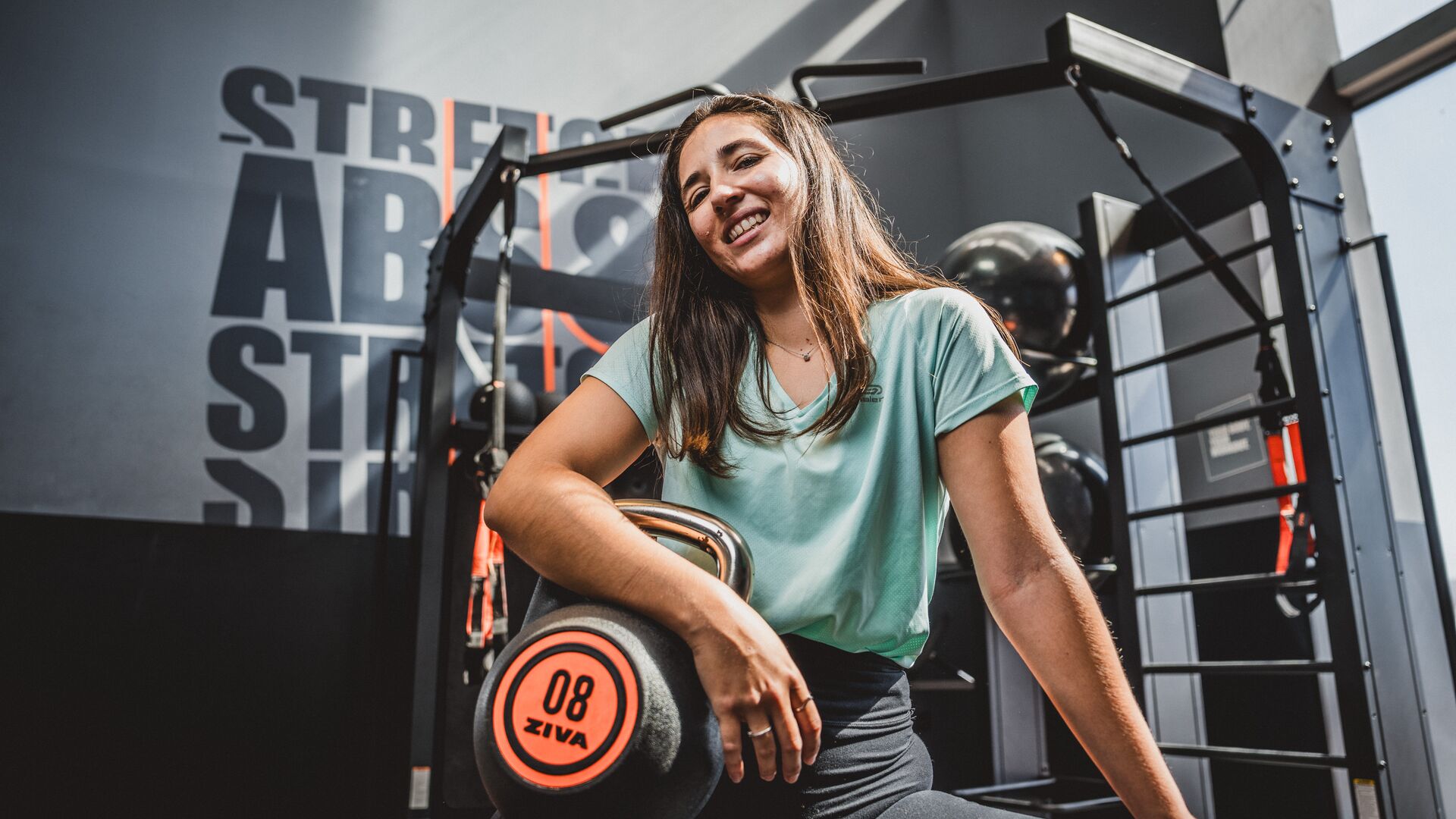 Woman in a mint green T-shirt sitting in the gym, leaning on an 8 kg kettlebell, smiling towards the camera.