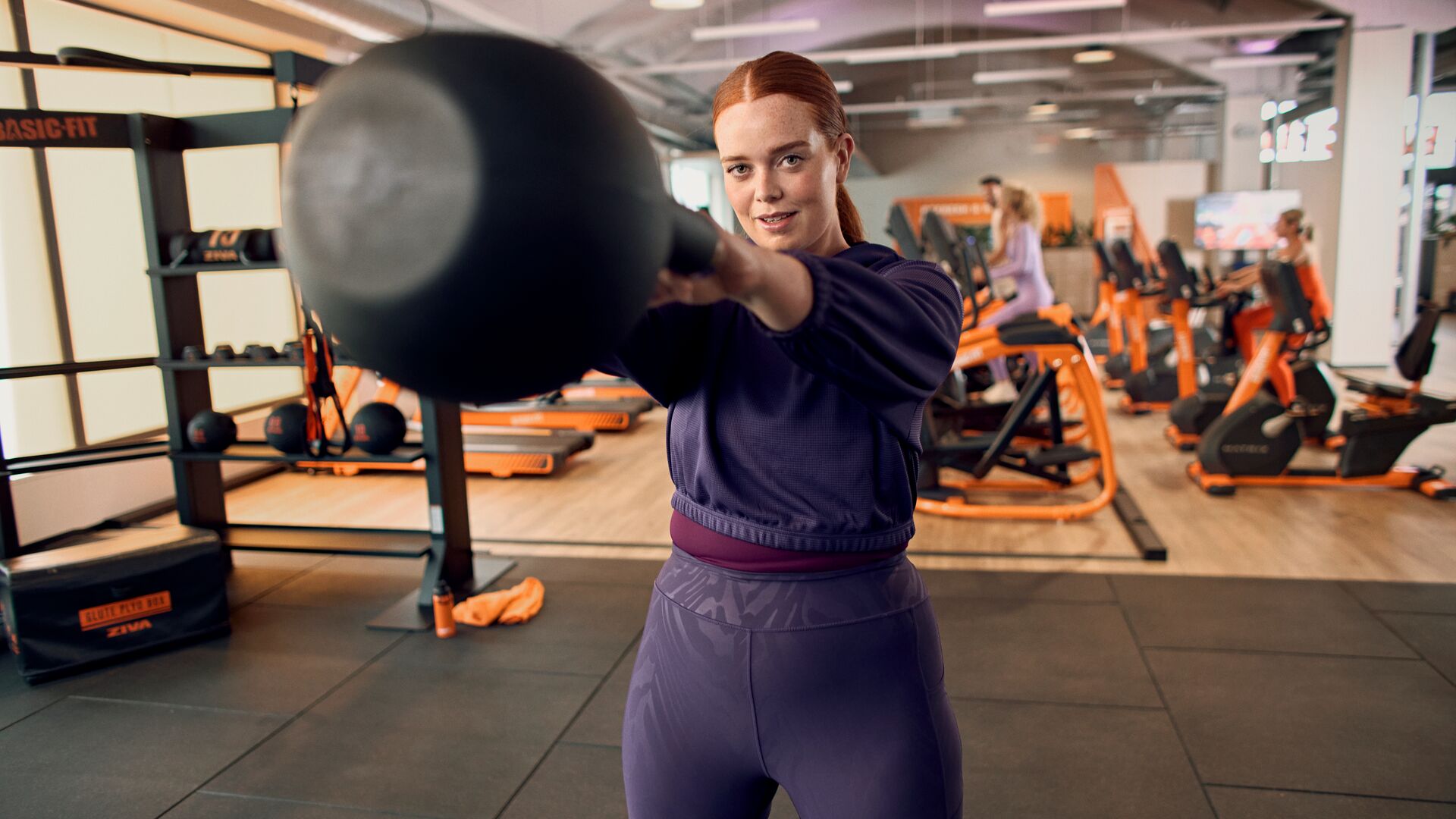 Woman training with a kettlebell in a Basic-Fit gym, holding the weight at shoulder height during a strength exercise.