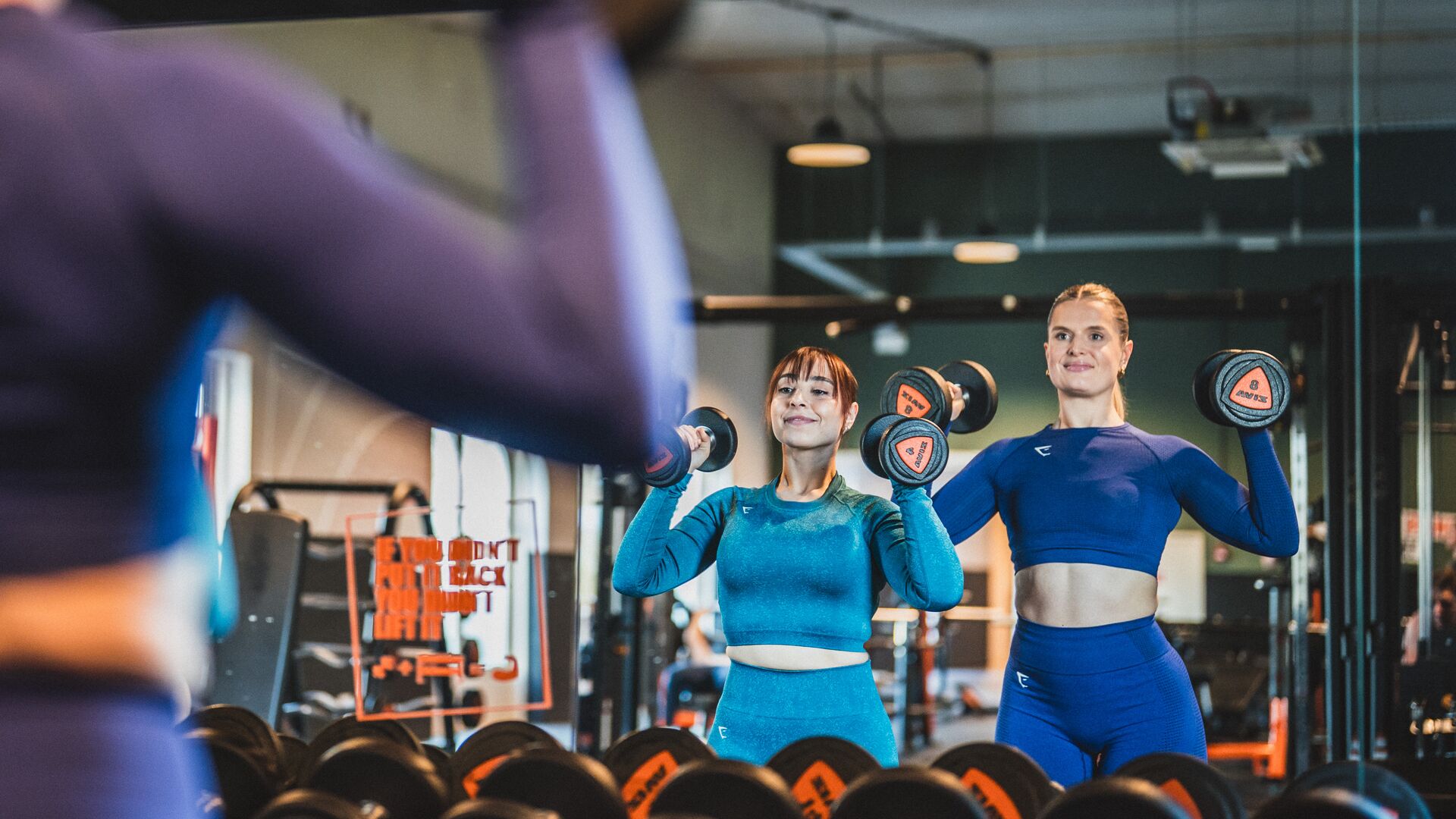 Two women lifting dumbbells in front of a mirror, focused and confident during their strength workout. They're dressed in colorful activewear, training side by side in a Basic-Fit gym. 