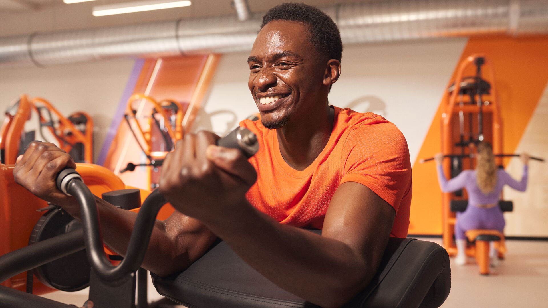 Person performing a seated bicep curl on a machine in a brightly lit gym, smiling during the exercise with other members training in the background.