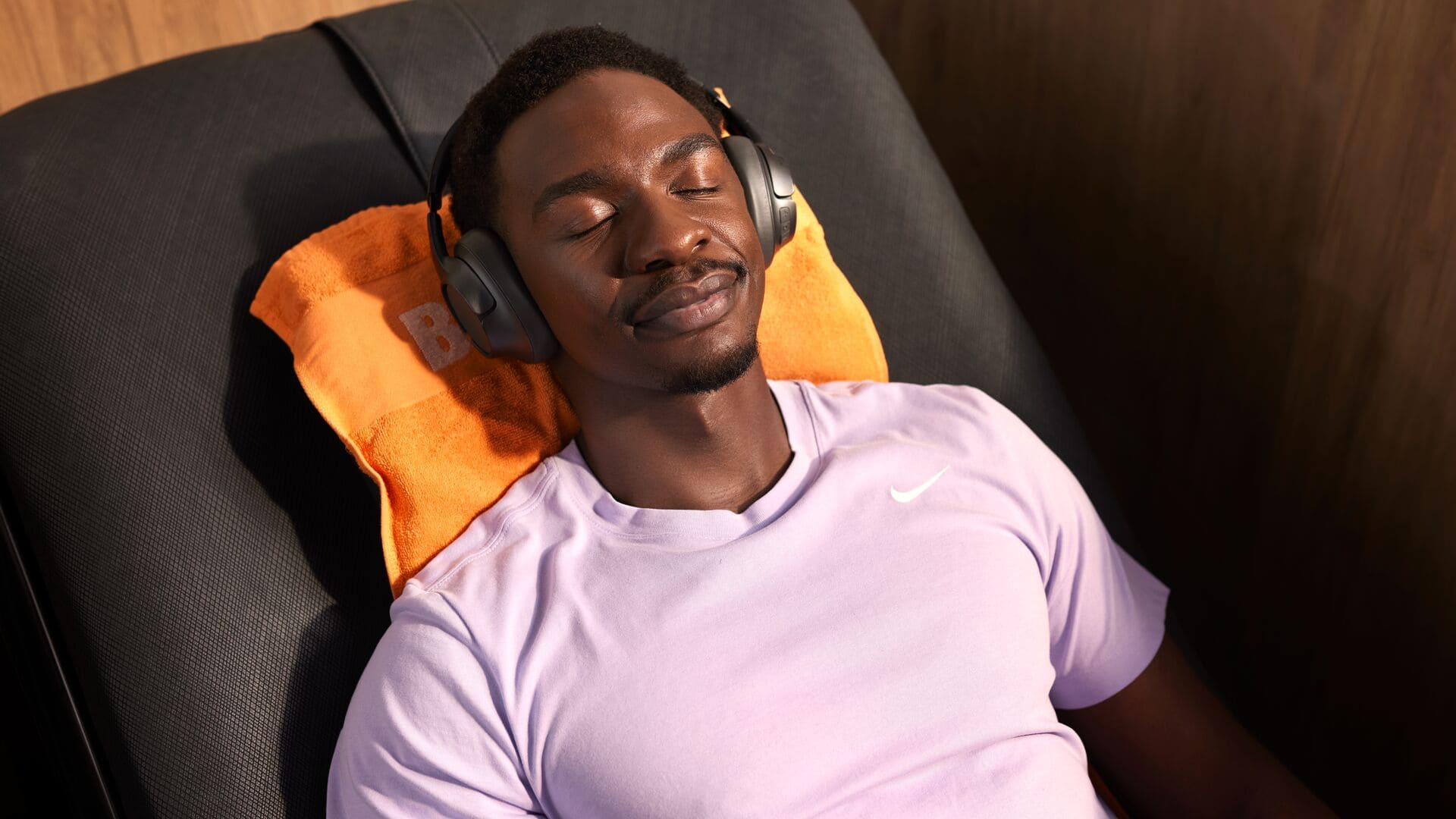 Ein Mann in einem hellvioletten Sportshirt entspannt mit Kopfhörern, liegt mit geschlossenen Augen auf einem orangefarbenen Handtuch in einem Basic-Fit-Massagesessel.