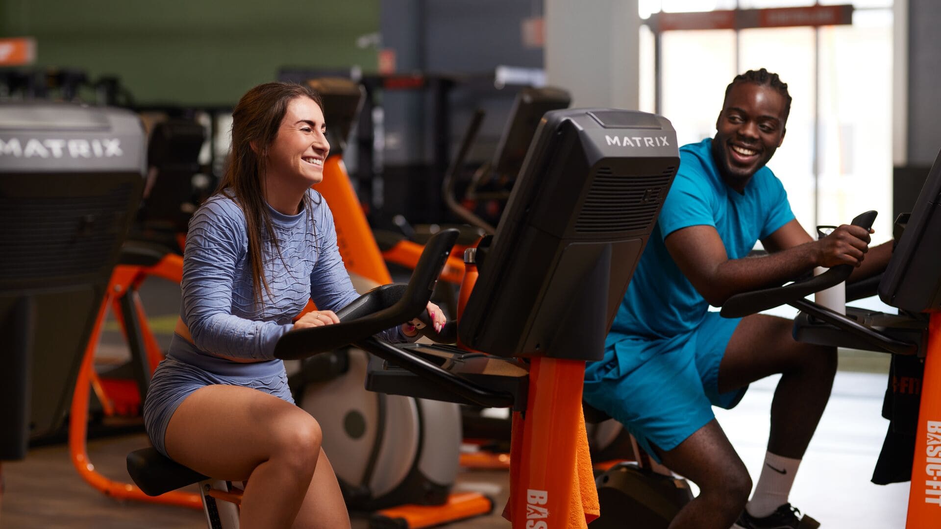 Eine Frau in hellblauem Sportset und ein Mann in türkisfarbener Kleidung sitzen auf Heimtrainern in einem Basic-Fit-Fitnessstudio, lächelnd und im Gespräch während ihres Cardiotrainings.