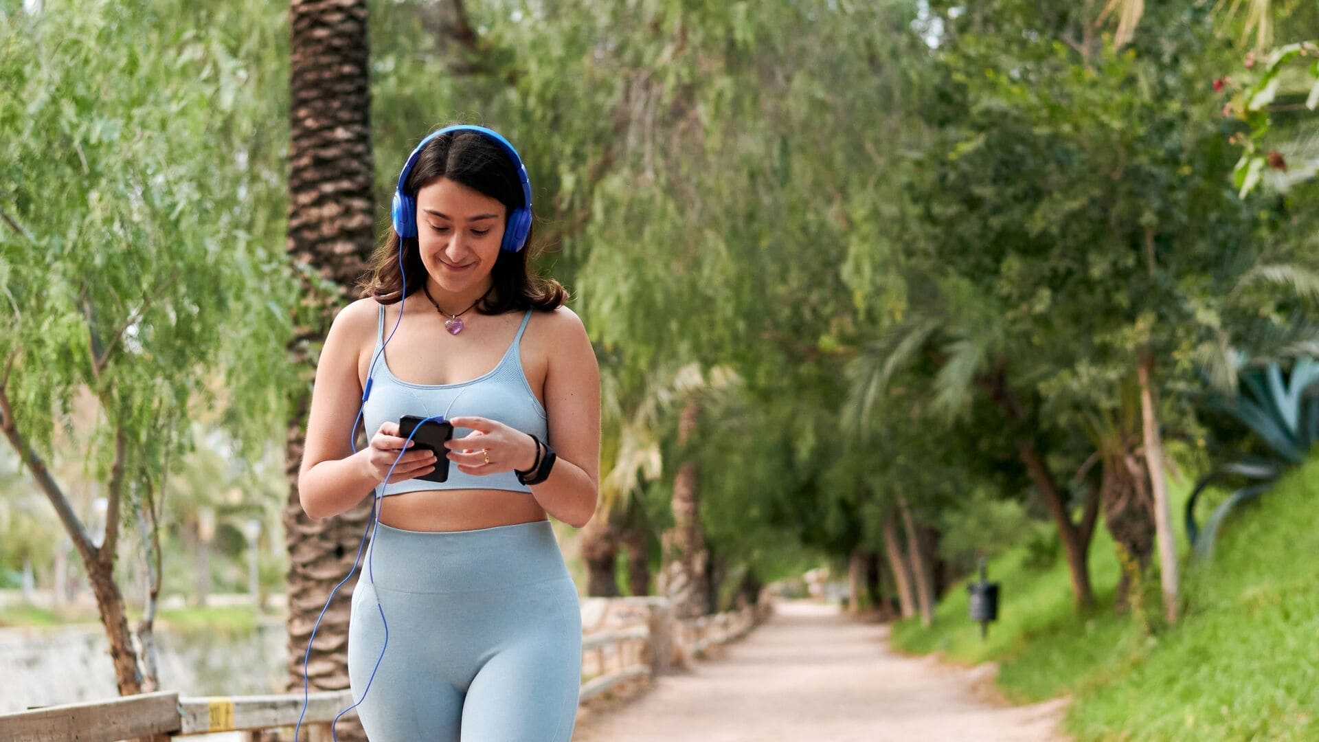 Een jonge vrouw in lichtblauwe sportkleding wandelt buiten op een met bomen omzoomd pad, met een koptelefoon op en haar telefoon in de hand. Ze ziet er ontspannen en gefocust uit tijdens haar wandeling of workout.