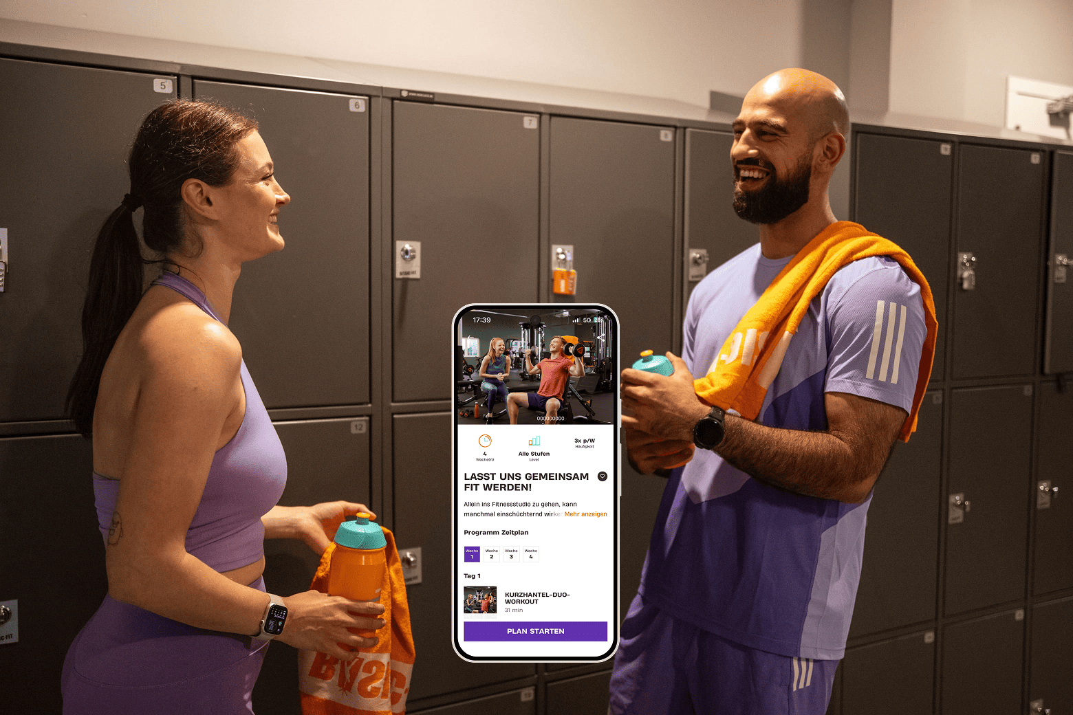 Zwei Personen stehen lachend in einem Fitnessstudio vor grauen Spinden und halten Trinkflaschen. In der Mitte ist ein Smartphone zu sehen, das ein Trainingsprogramm mit dem Titel „Lasst uns gemeinsam fit werden!“ auf der Basic-Fit App zeigt.