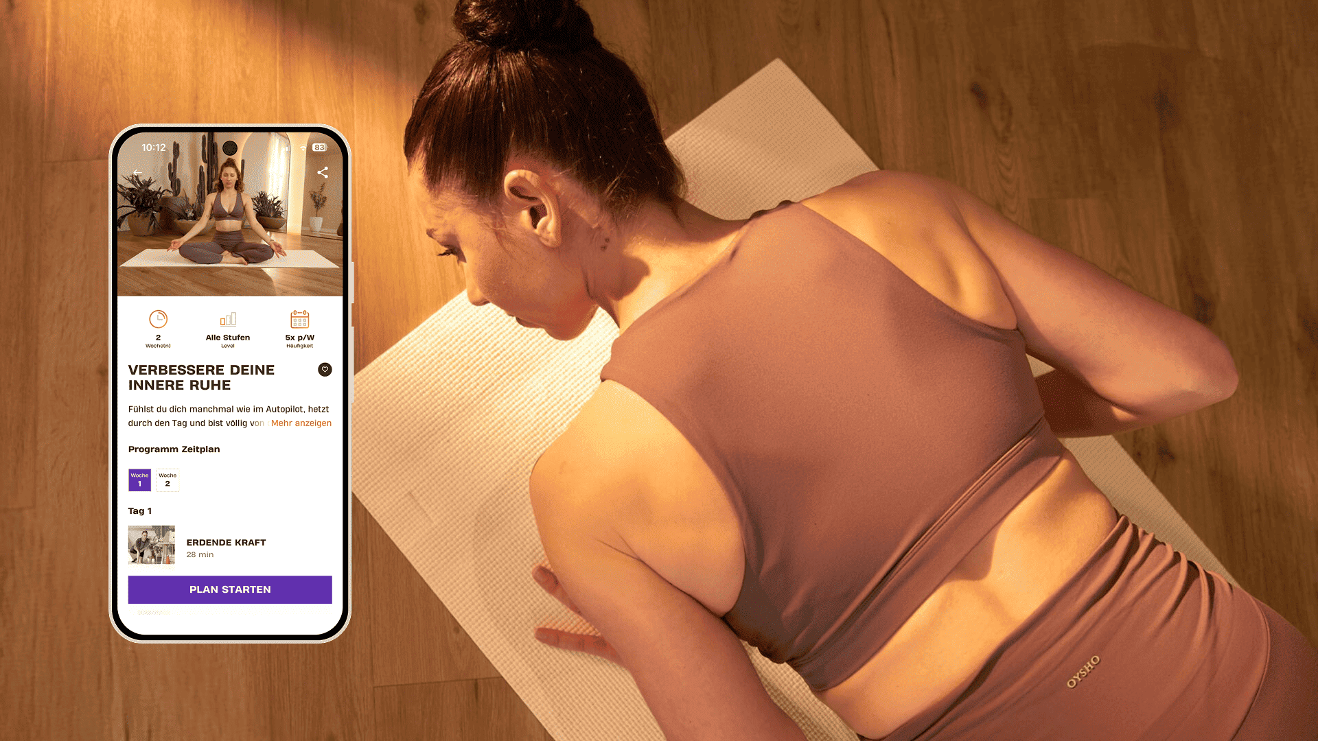 Frau in mauvefarbener Sportkleidung liegt auf einer Yogamatte in warm beleuchtetem Raum mit Holzboden, neben einem Smartphone mit dem Basic-Fit-Programm „Verbessere deine innere Ruhe“.