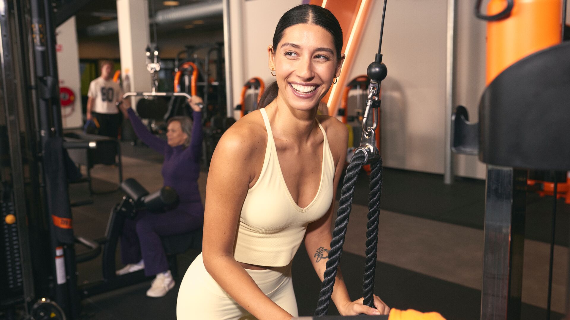 Een vrouw glimlacht terwijl ze een kabeltoestel gebruikt in een Basic-Fit club en een touwgreep vasthoudt tijdens een krachtoefening. Andere sporters trainen op de achtergrond.