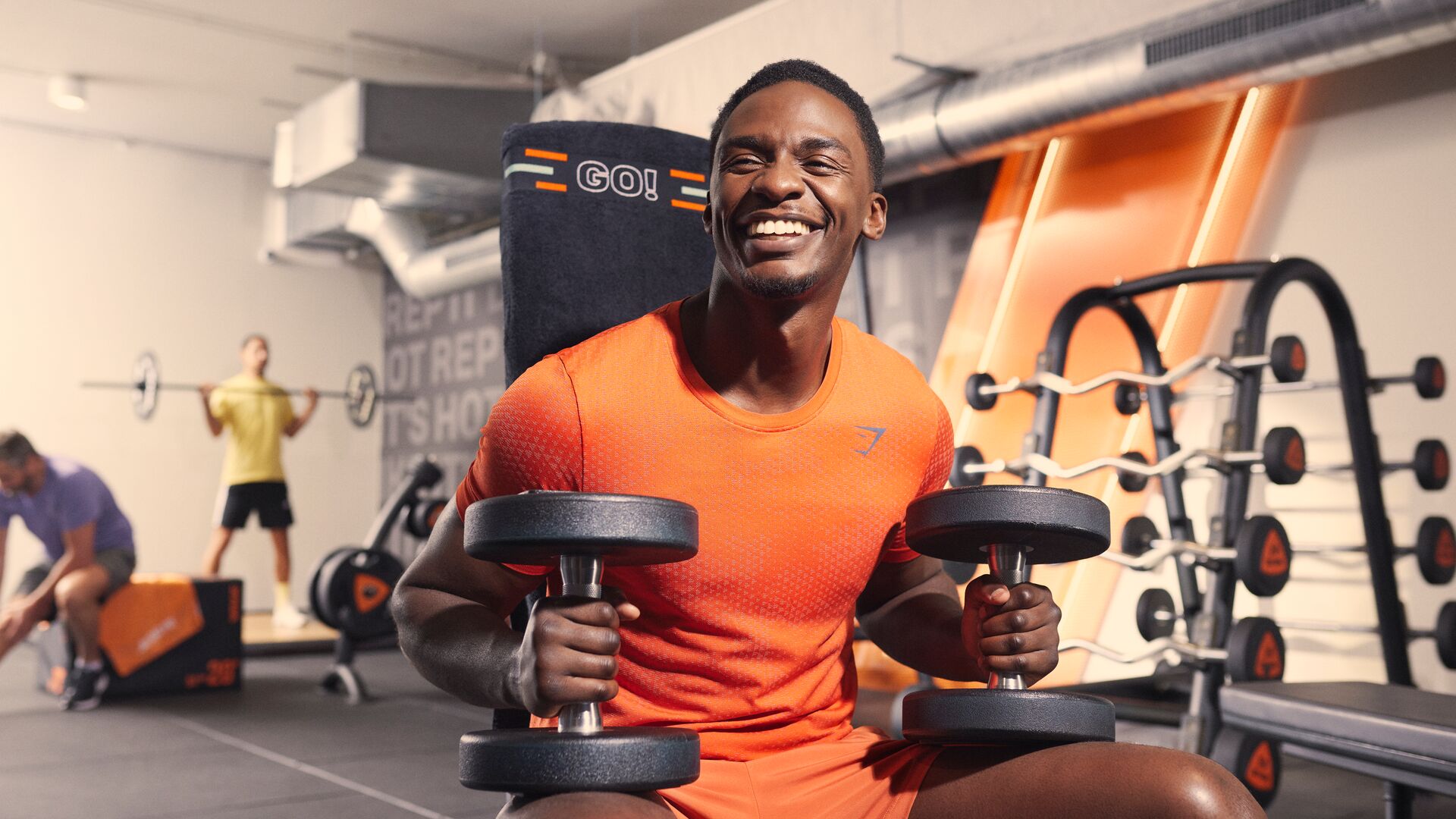 Een man zit glimlachend op een bankje in een Basic-Fit club en houdt twee dumbbells vast tijdens een krachttraining. Andere sporters trainen op de achtergrond tussen krachttoestellen en oranje accenten.